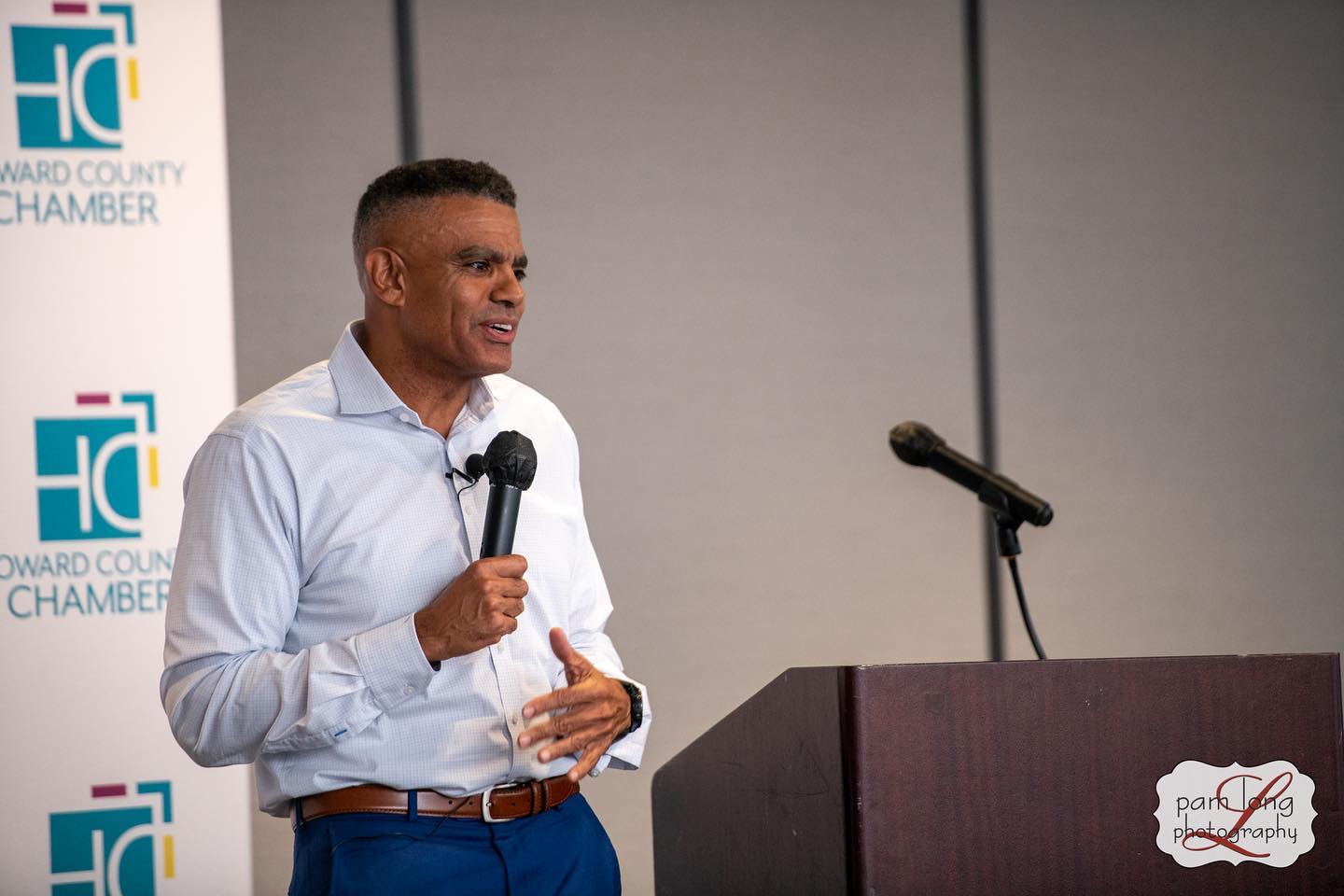 A man in a light blue shirt speaks into a microphone at a podium. In the background, a banner for Howard County Chamber is partially visible. The podium features a photography business logo, attracting job seekers and boosting social capital smart initiatives.
