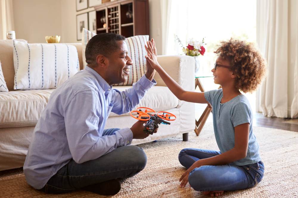 A man and a young girl are sitting on the floor in a living room, smiling and giving each other a high five. The man is holding a small toy drone. They appear to be having a playful moment together.