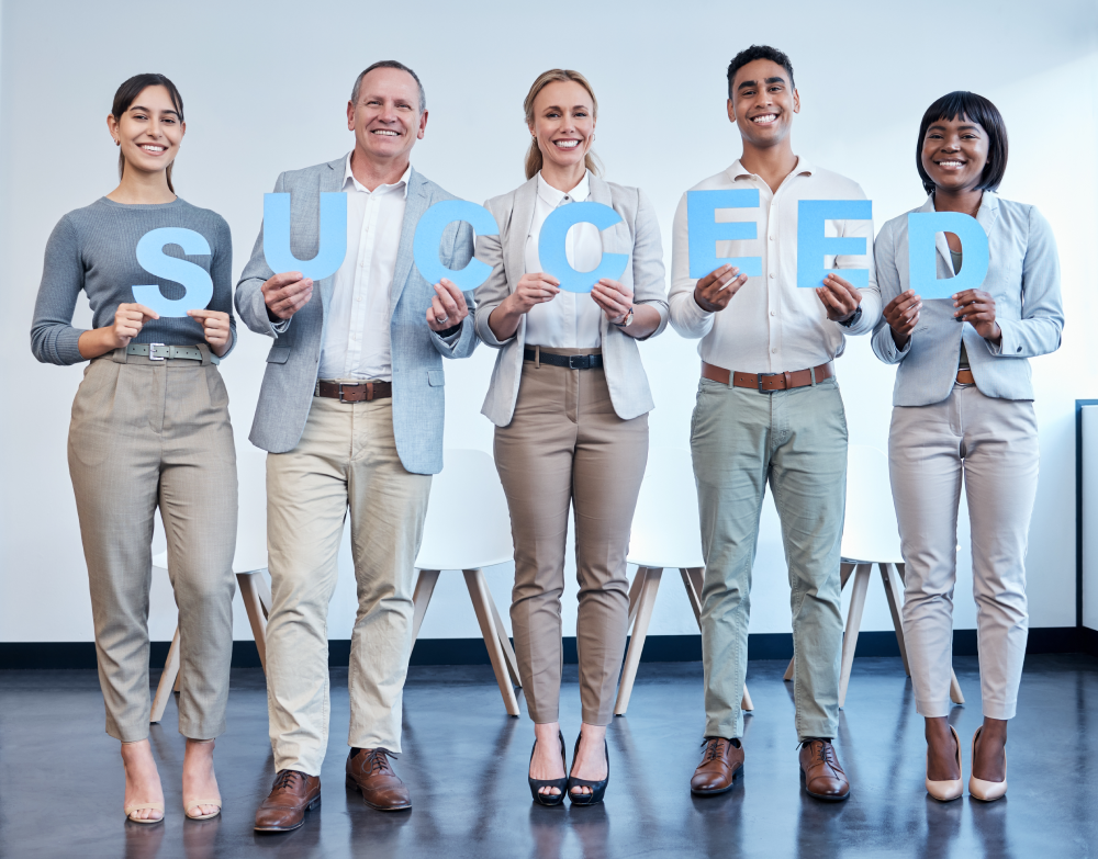 A group of five diverse business professionals smiling and holding blue letters that spell SUCCEED in a bright, modern office setting. They are dressed in business attire, standing in a line against a white wall.