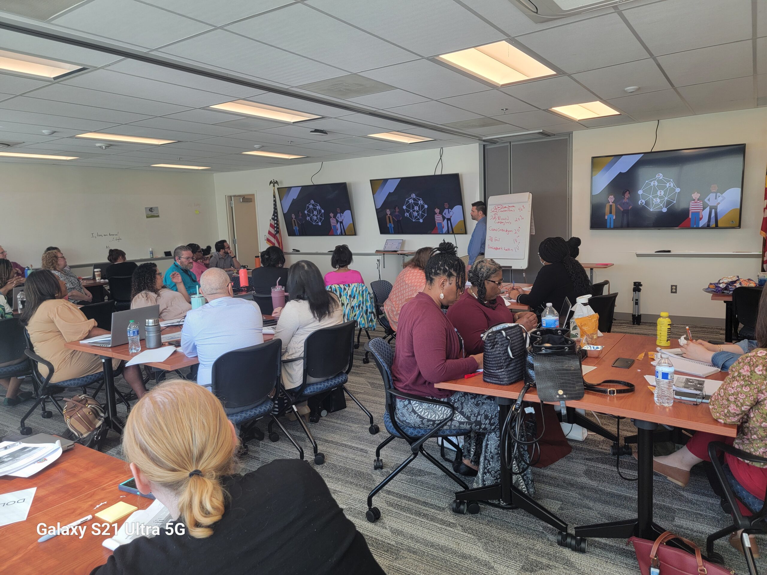 A classroom with people seated at tables, watching a presentation on three large screens. Attendees are taking notes and engaging in discussion. An American flag and a whiteboard with notes are visible near the front.