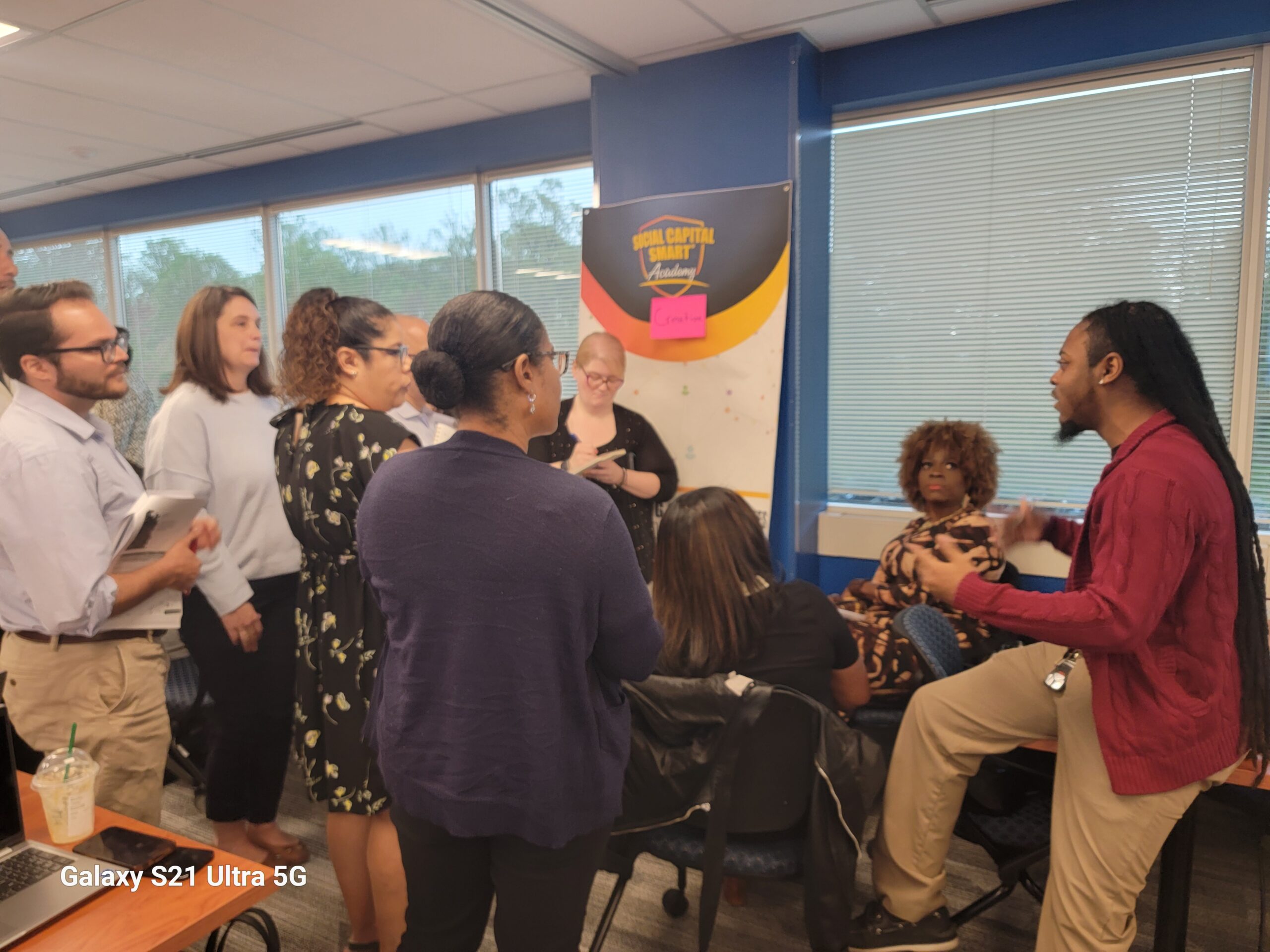 A group of people having a discussion in an office, with a man seated and gesturing while others stand and listen. A Social Capital Launch poster is visible in the background.
