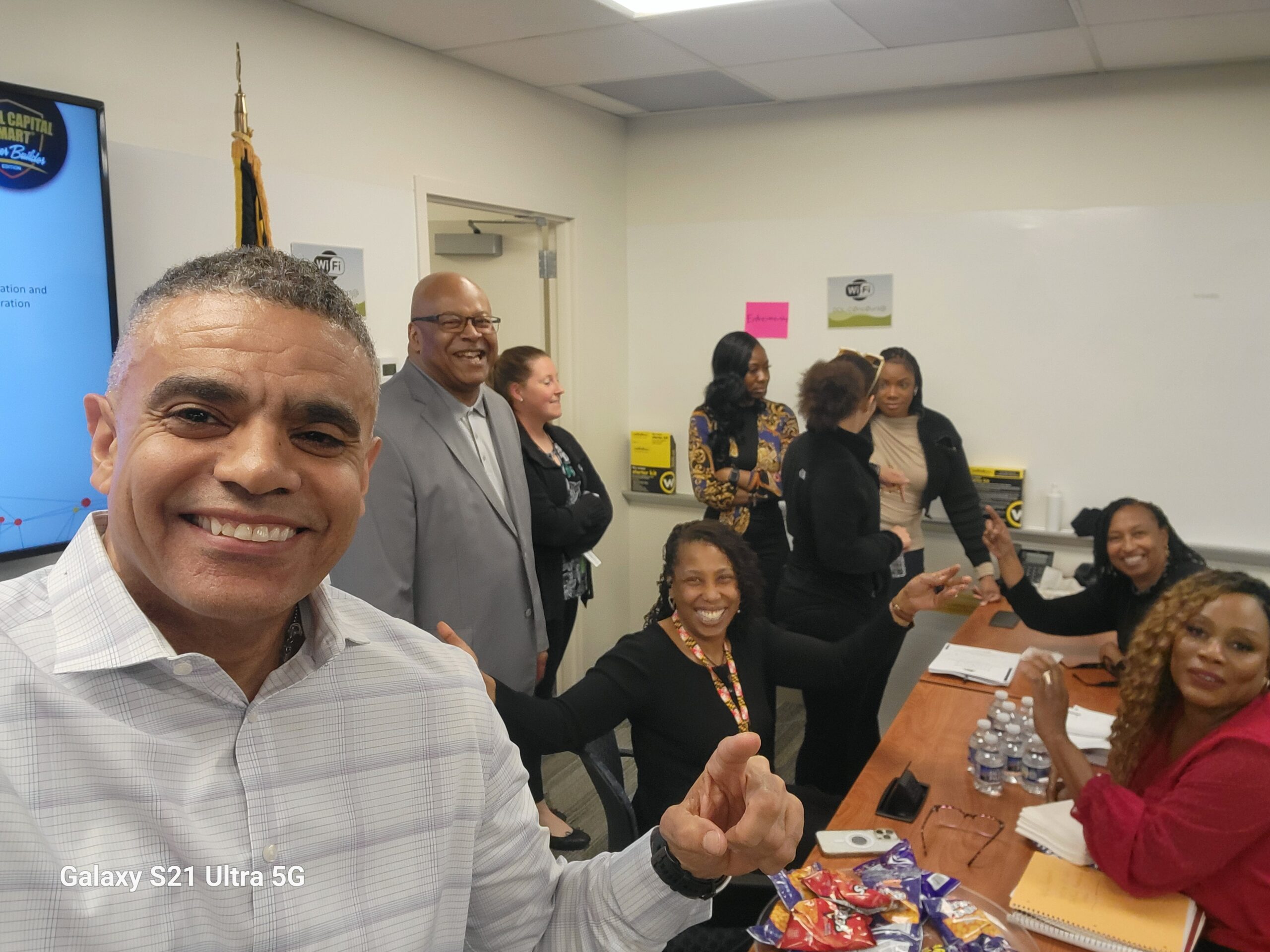 A group of people smiling and posing at a meeting room table. One person in the foreground is taking a selfie. Others are seated and standing, engaged in conversation. There are snacks and water bottles on the table.