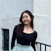 A person with long dark hair smiles while standing outdoors. They are wearing a black long-sleeve top and a patterned skirt. The background includes a white textured wall.